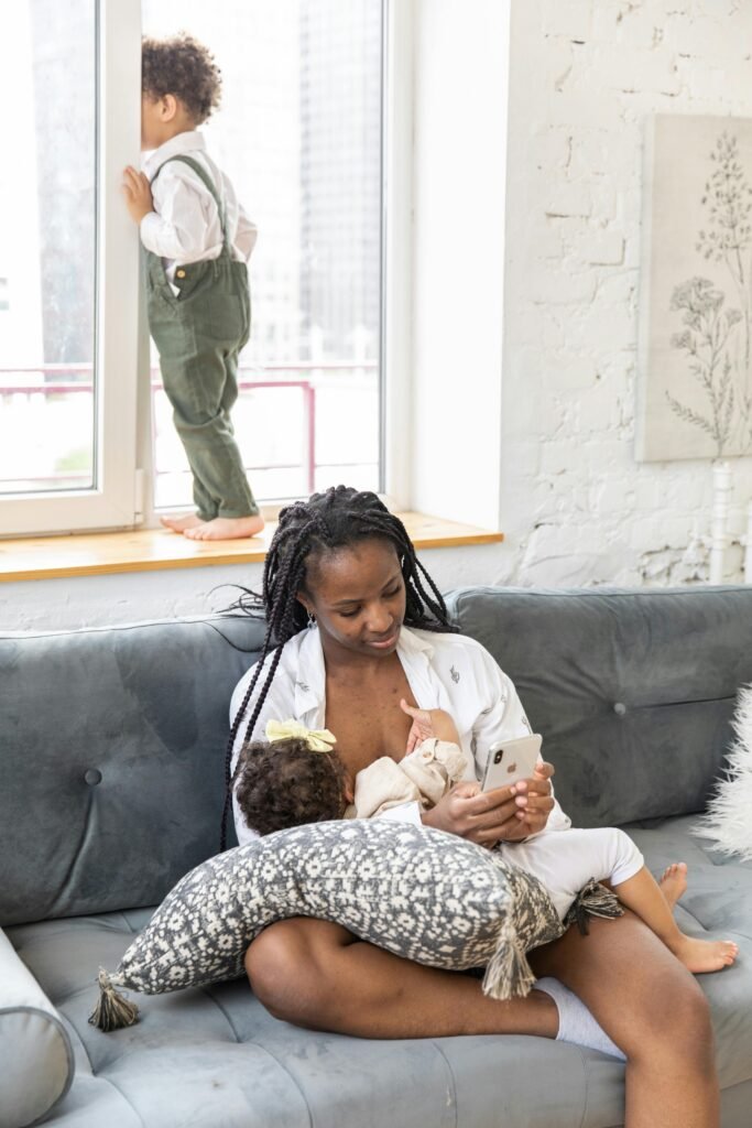 A mother breastfeeds her baby on a couch while using her smartphone, with another child looking out the window.